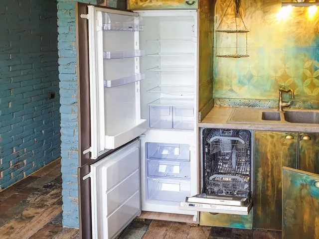 Frigorífico abierto y vacío junto a un lavavajillas en una cocina con azulejos dorados y pared de ladrillo
