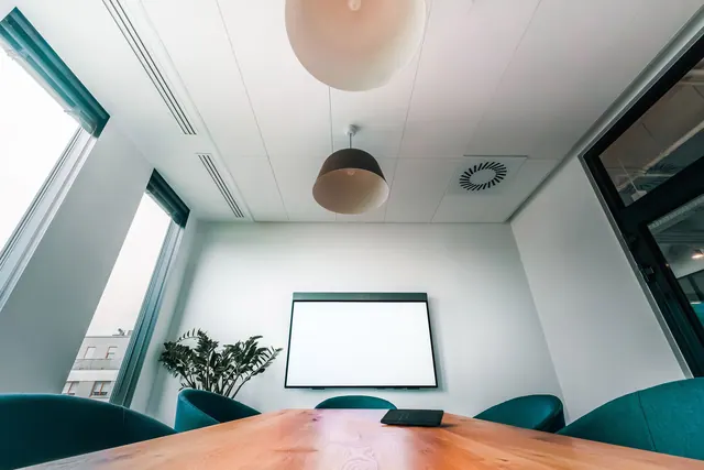 Sala de reuniones pequeña con una pantalla grande para videoconferencia y varias personas alrededor de la mesa