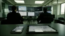 Dos personas en una sala de control con múltiples pantallas de monitoreo