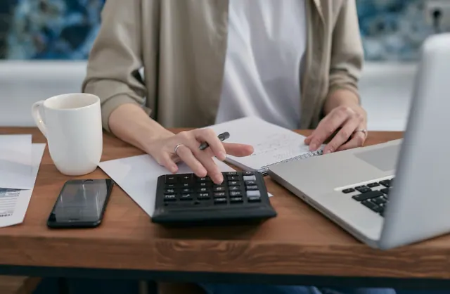 Una persona usa una calculadora junto a un portátil y un cuaderno