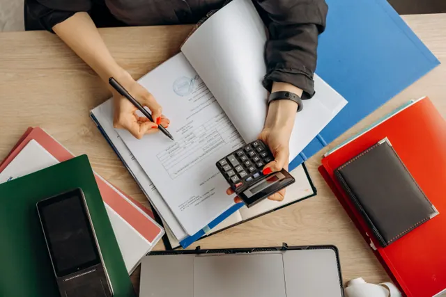 Una persona calcula cifras con documentos y carpetas sobre una mesa