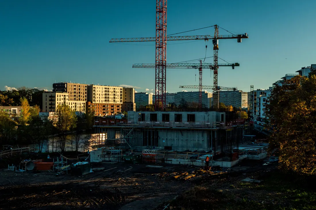 Obra en construcción con grúas junto a un río, rodeada de bloques de viviendas