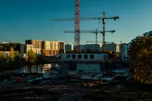 Obra en construcción con grúas junto a un río, rodeada de bloques de viviendas