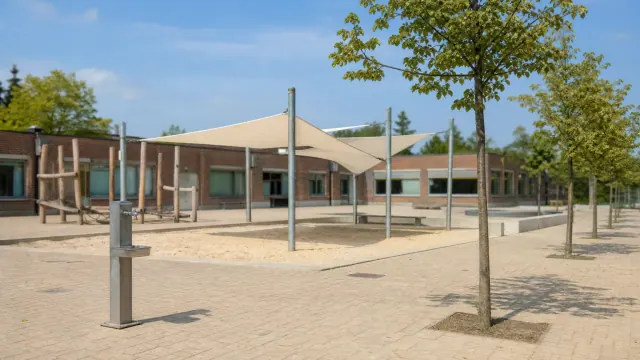 Patio de arena con toldo de lona beige, árboles jóvenes y edificio de ladrillo con ventanas al fondo bajo cielo