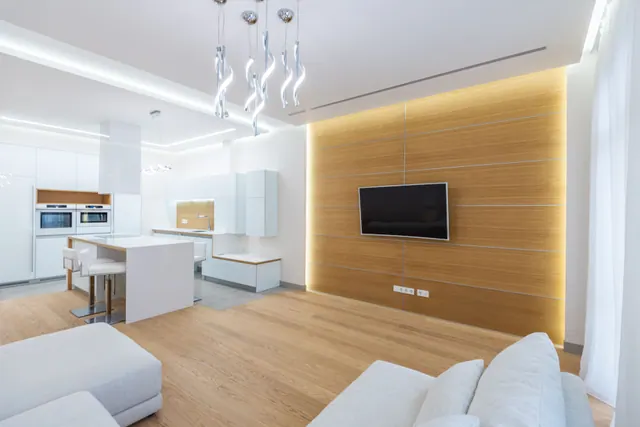 Salón moderno con paredes de madera clara, televisor plano empotrado, sofá blanco, suelo de madera y cocina integrada