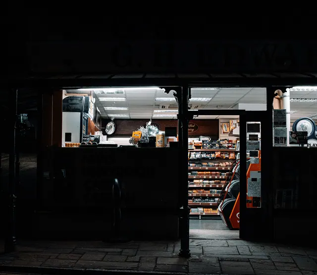 Fachada de un pequeño comercio cerrado con escaparates iluminados y estanterías con productos en el interior