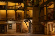 Patio interior nocturno de arquitectura medieval con galerías de madera, balcones y fachadas de piedra iluminadas