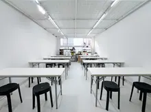 Espacio diáfano con mesas plegables blancas y taburetes negros, techo de rejilla con iluminación LED y zona de trabajo