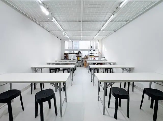 Espacio diáfano con mesas plegables blancas y taburetes negros, techo de rejilla con iluminación LED y zona de trabajo