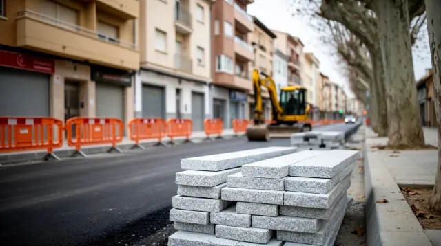 Pila de adoquines de hormigón gris en primer plano junto a una calle en obras con maquinaria amarilla y barrera naranja