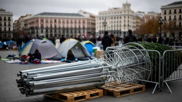 Vallas y tubos metálicos apilados en una plaza urbana con tiendas de campaña al fondo