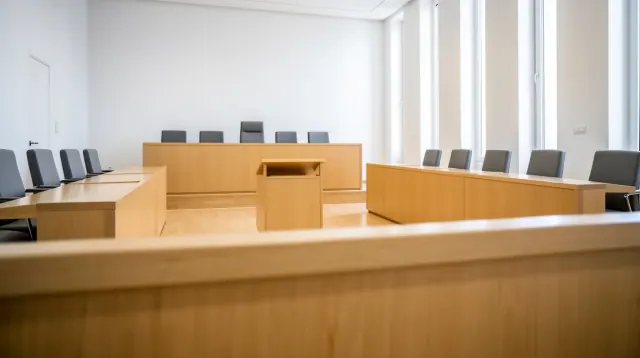 Sala de vistas con mobiliario de madera clara, tribunal elevado al fondo y sillas grises dispuestas en los laterales