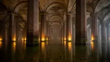Sótano subterráneo con columnas de ladrillo y arcos de piedra iluminados, suelo inundado que refleja la luz amarilla