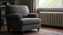Sillón tapizado en gris oscuro con brazos redondeados en una sala iluminada por ventanal, junto a radiador blanco