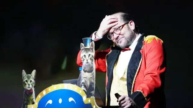 Un humorista con levita roja de maestro de ceremonias se lleva la mano a la frente junto a recortes de gatos