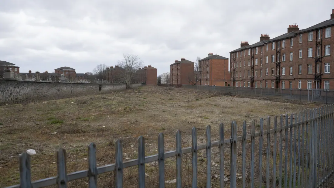 Terreno vacío cercado con valla metálica junto a edificios residenciales de ladrillo