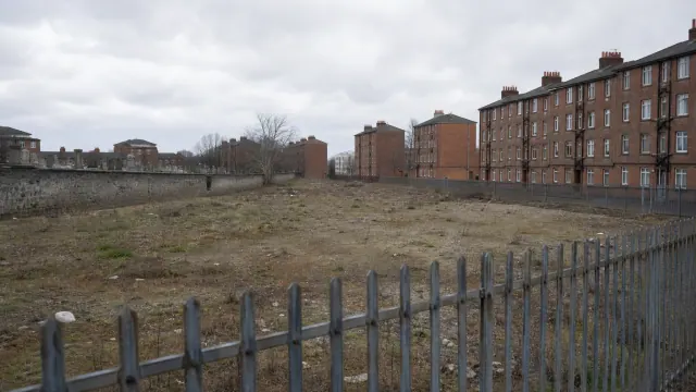 Terreno vacío cercado con valla metálica junto a edificios residenciales de ladrillo