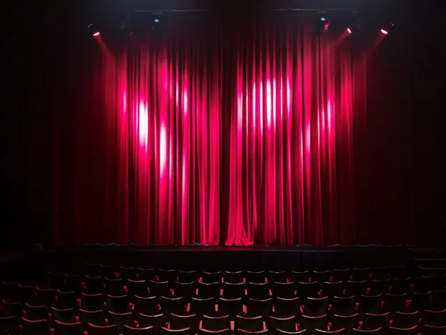 Escenario de un teatro con el telón rojo iluminado y butacas vacías en primer plano