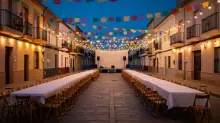 Calle engalanada con banderines de colores y mesas largas con mantel blanco frente a un pequeño escenario