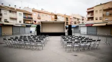 Sillas metálicas grises en filas frente a un escenario montado en una plaza con puestos cerrados