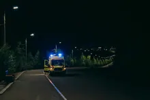 Ambulancia con luz azul activada circulando por una carretera rural nocturna entre árboles y farolas