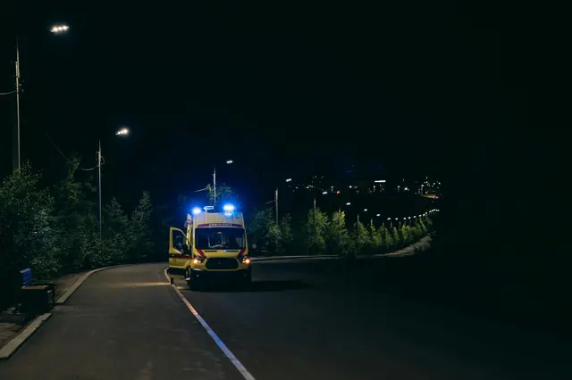 Ambulancia con luz azul activada circulando por una carretera rural nocturna entre árboles y farolas