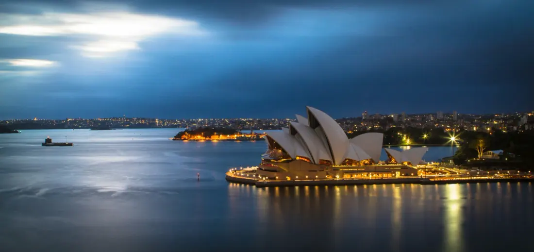 La Ópera de Sídney iluminada al anochecer junto a la bahía