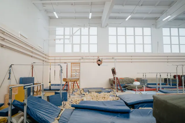 Sala de rehabilitación vacía con barras paralelas, camilla y material de equilibrio junto a una ventana