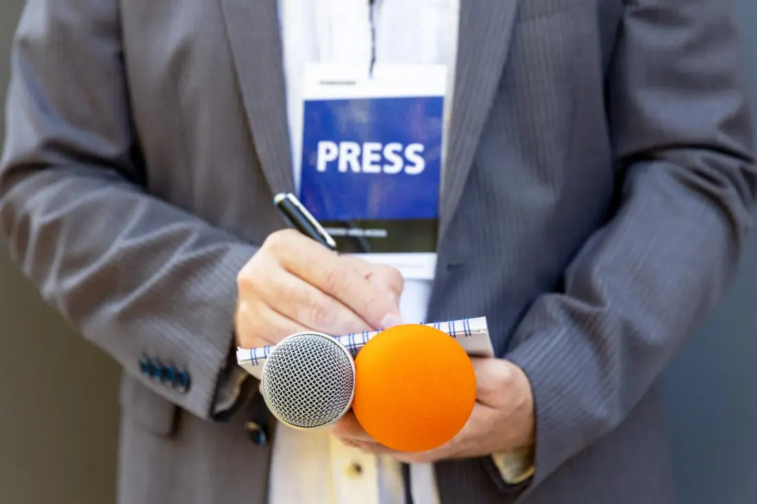 Periodista con acreditación de prensa sostiene dos micrófonos y un cuaderno