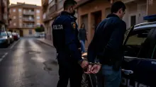 Dos policías escoltan a una persona esposada junto a un vehículo en una calle urbana