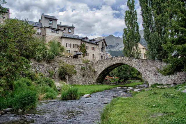 Pueblo medieval junto a un río con puente de piedra, edificios de piedra y montañas al fondo bajo cielo nublado